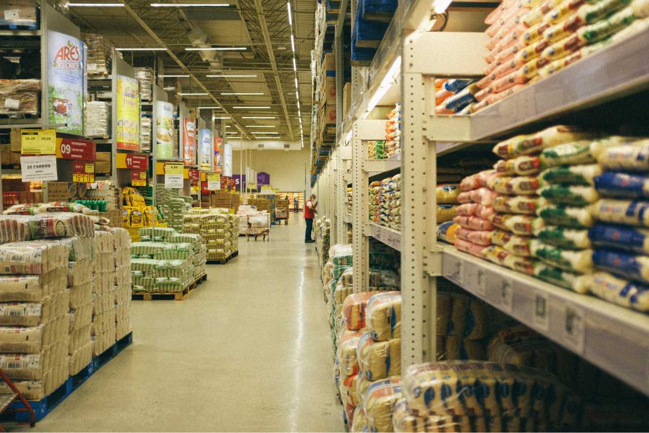 A deserted aisle in a large wholesale store with one worker checking the shelves.