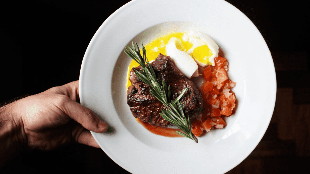 Hand holding a white plate of meat eggs and tomatoes